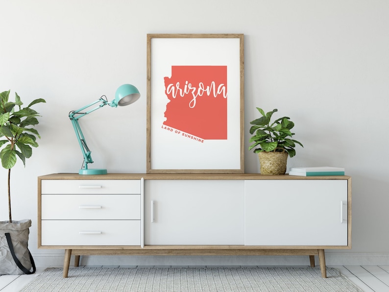 Op de afbeelding: Een ingelijste kunstdruk met de staat Arizona in koraal, met het woord "arizona" in wit script. De tekst "LAND OF SUNSHINE" staat onder de omtrek van de staat. De print staat op een witte en houten kast, met een turquoise bureaulamp en potplanten. De kast is 150 cm breed.