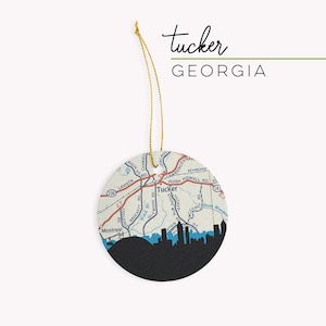May include: A round, white ornament with a map of Tucker, Georgia, and a black silhouette of the city skyline. The ornament has a gold string for hanging.