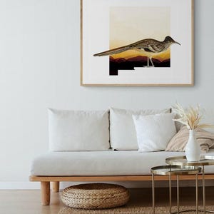 May include: A framed art print featuring a roadrunner bird illustration. The bird is depicted with brown and white feathers, standing against a backdrop of a sunset-colored landscape. The artwork is encased in a light-colored wooden frame, suitable for home decor.