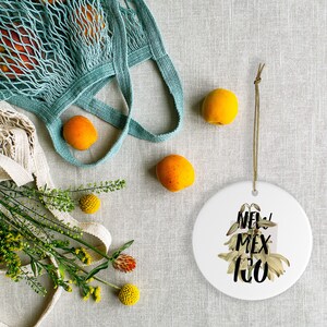 May include: A white ceramic ornament with the text "NEW MEX 100" and floral accents hangs by a brown string. Apricots, a teal net bag, and yellow flowers are scattered on a neutral linen surface.