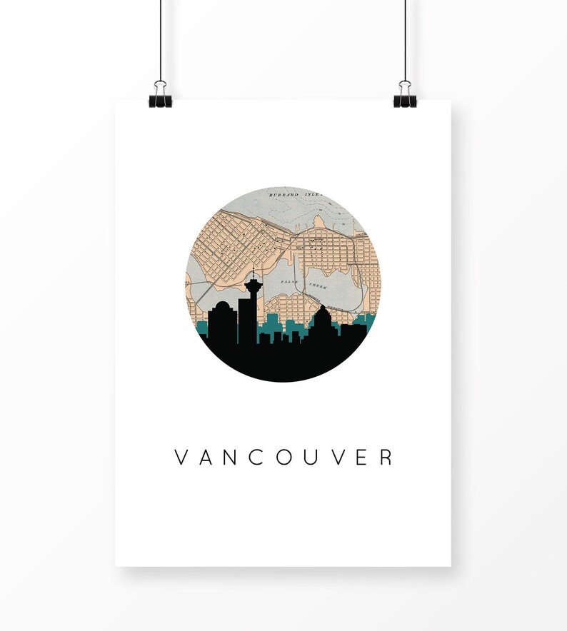 Vancouver art print hanging from two black clips against a plain white background, featuring a circular map and silhouette skyline above the text VANCOUVER.