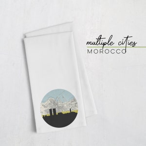 May include: White kitchen towel with a circular graphic featuring a city skyline in black, yellow, and blue. The text "multiple cities MOROCCO" is printed in black and green.