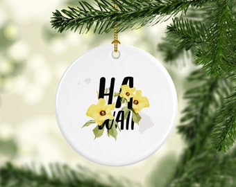 May include: White ceramic ornament with the word "HAWAII" in black lettering and yellow floral accents. The ornament is round and has a gold string for hanging. The background is blurred, suggesting a festive setting.