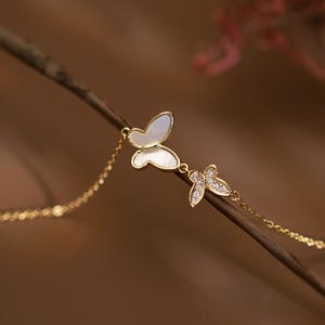 May include: A delicate gold-colored necklace featuring two butterfly pendants. One butterfly has a white, iridescent surface, while the other is adorned with small, sparkling crystals. The necklace is displayed on a thin, brown branch against a blurred background.