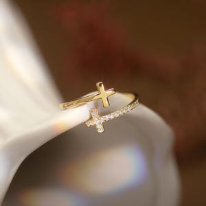 Può includere: Un anello dorato con due motivi a croce. Una croce è semplice, mentre l'altra è incastonata con piccole pietre preziose trasparenti. L'anello è posto su uno sfondo morbido e sfocato, evidenziando i dettagli del gioiello.