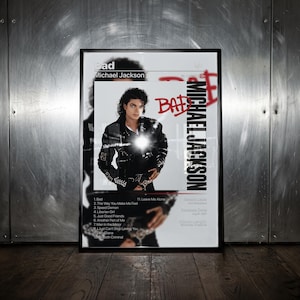 Może przedstawiać: Oprawiony plakat z okładką albumu Bad Michaela Jacksona. Projekt zawiera czarno-białe zdjęcie artysty, czerwony napis Bad oraz listę utworów. Plakat znajduje się w czarnej ramie na tle metalowej ściany.