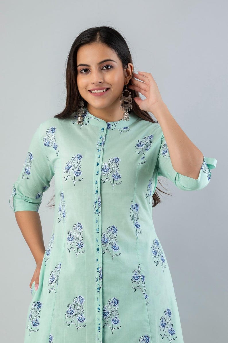 May include: A light green, button-down dress with a floral pattern in blue and white. The dress has a mandarin collar and three-quarter sleeves. The model is wearing silver earrings.