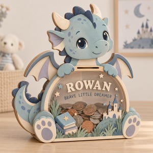 May include: A wooden money box shaped like a light blue dragon with a clear front panel. The text ROWAN and BRAVE LITTLE DREAMER is printed on the front. Inside the box are various metal coins, a small castle, and two books.