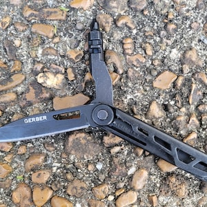 May include: A black multi-tool with the brand name "GERBER" visible on the blade. The tool is open, displaying a knife blade and other implements. The tool is lying on a surface of small rocks and pebbles.