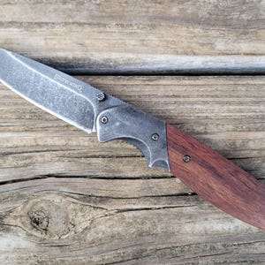 May include: A folding knife with a silver blade and a wooden handle. The word "BROWNING" is etched on the blade. The handle is a dark brown color and is attached to the blade with silver rivets. The knife is resting on a wooden surface.