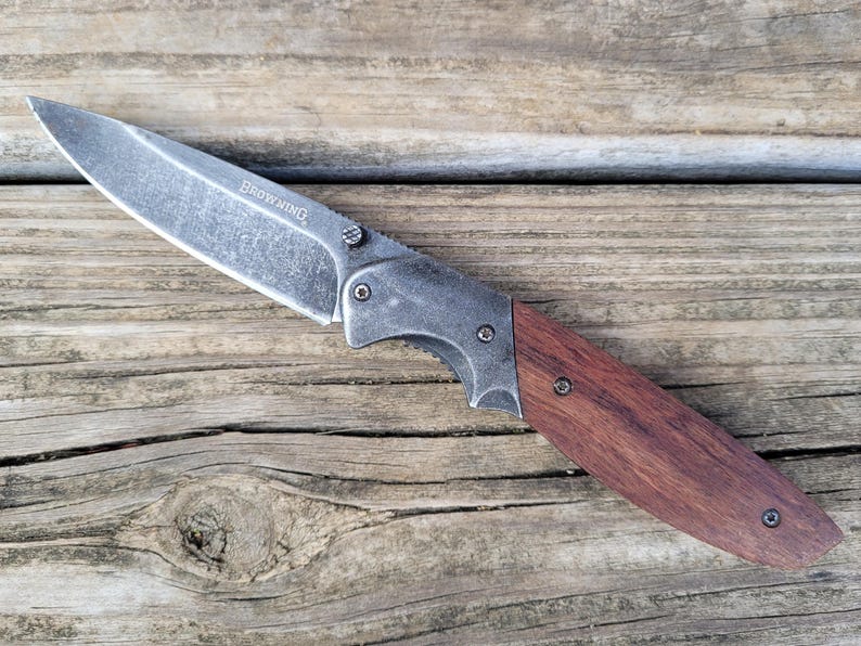 May include: A folding knife with a silver blade and a wooden handle. The word "BROWNING" is etched on the blade. The handle is a dark brown color and is attached to the blade with silver rivets. The knife is resting on a wooden surface.