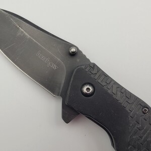 Op de afbeelding: Een zwart vouwmes met een gedeeltelijk gekarteld lemmet. Het lemmet heeft de merknaam "Kershaw" gegraveerd. De handgreep is zwart met een textuurpatroon. Het mes is ontworpen voor dagelijks gebruik en buitengebruik.