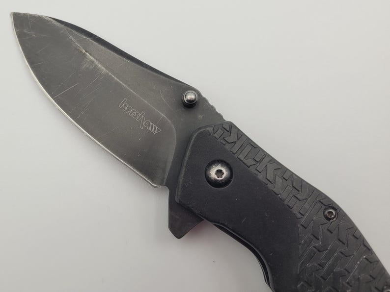 Op de afbeelding: Een zwart vouwmes met een gedeeltelijk gekarteld lemmet. Het lemmet heeft de merknaam "Kershaw" gegraveerd. De handgreep is zwart met een textuurpatroon. Het mes is ontworpen voor dagelijks gebruik en buitengebruik.