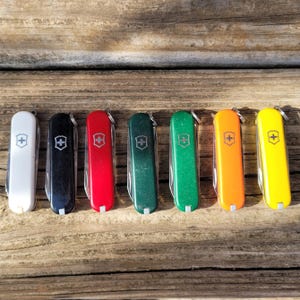 May include: A collection of seven colorful Swiss Army knives arranged on a weathered wooden surface. The knives come in white, black, red, two shades of green, orange, and yellow. Each knife features a silver emblem and a small keyring.