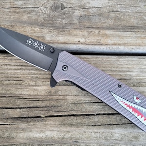 May include: A gray folding knife with a black blade. The blade has three starburst designs. The handle is gray with a textured pattern and a shark mouth design in white, red, and black. The knife is on a wooden surface.