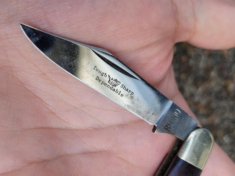 Może przedstawiać: Składany scyzoryk ze stali nierdzewnej z częściowo otwartym ostrzem. Ostrze ma wygrawerowane słowa "Tough Sharp Dependable" i logo nosorożca. Rękojeść jest ciemnobrązowa, a n&oacute;ż jest oznaczony napisem "RHINO".