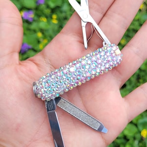 May include: A small, multi-tool with a bedazzled handle. The handle is covered in iridescent rhinestones. The tool includes a small knife, nail file, and scissors. The blades and scissors are silver-colored.