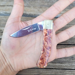 May include: A small, open pocket knife with a polished steel blade and a handle featuring a swirling brown and cream pattern. The blade is marked with the text "Elk Ridge". The knife is open, revealing the sharp edge.