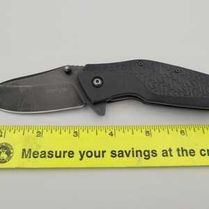 Op de afbeelding: Een zwart vouwmes met een getextureerd handvat en een gedeeltelijk gekarteld lemmet. Op het lemmet staat de merknaam "Kershaw" gedrukt. Onder het mes ligt een gele liniaal.