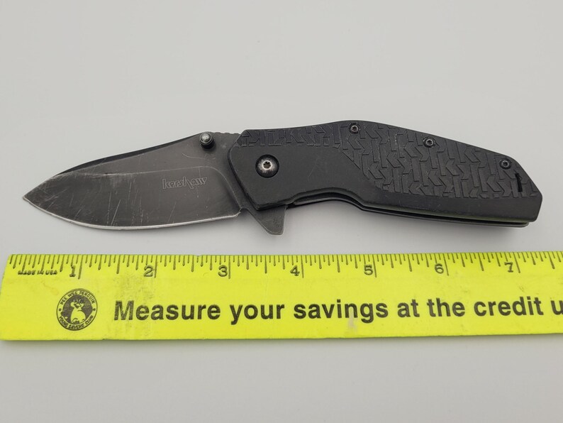 Op de afbeelding: Een zwart vouwmes met een getextureerd handvat en een gedeeltelijk gekarteld lemmet. Op het lemmet staat de merknaam "Kershaw" gedrukt. Onder het mes ligt een gele liniaal.