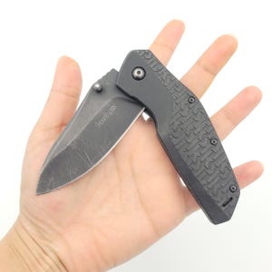 Op de afbeelding: Een zwart vouwmes met een gedeeltelijk open lemmet. Het lemmet heeft een versleten uiterlijk. De handgreep is zwart met een textuurpatroon. De merknaam "Kershaw" is op het lemmet gedrukt.