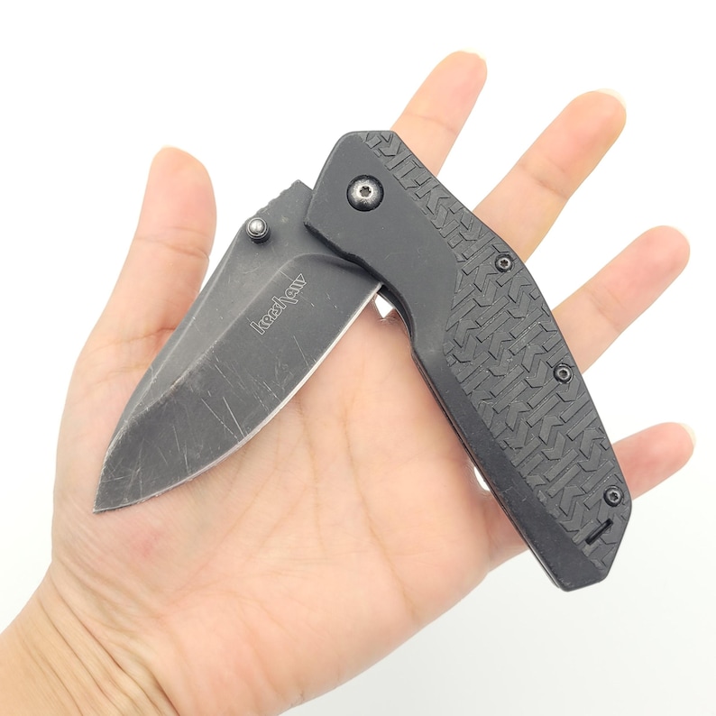 Op de afbeelding: Een zwart vouwmes met een gedeeltelijk open lemmet. Het lemmet heeft een versleten uiterlijk. De handgreep is zwart met een textuurpatroon. De merknaam "Kershaw" is op het lemmet gedrukt.