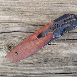 May include: A folding pocket knife with a wooden handle and a metal clip. The blade is partially visible, and the knife is set against a weathered wooden surface. The knife has a brown handle and a silver blade.