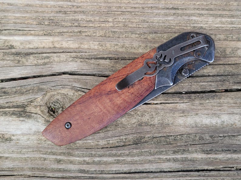 May include: A folding pocket knife with a wooden handle and a metal clip. The blade is partially visible, and the knife is set against a weathered wooden surface. The knife has a brown handle and a silver blade.