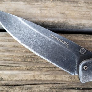May include: Close-up of a Browning folding knife with a weathered, gray blade. The word "BROWNING" is etched on the blade. The knife is set against a wooden background.