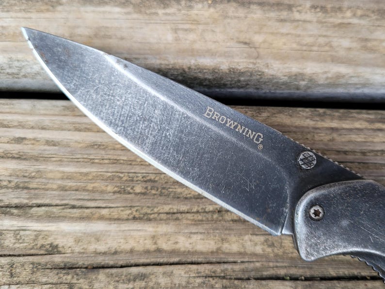 May include: Close-up of a Browning folding knife with a weathered, gray blade. The word "BROWNING" is etched on the blade. The knife is set against a wooden background.