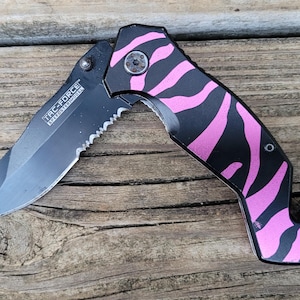 May include: A black and pink folding knife with a zebra stripe pattern. The blade is partially serrated and has the text "TAC-FORCE SPEEDSTER MODEL". The knife is open, revealing the blade and handle.