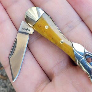 Op de afbeelding: Een klein, open zakmes met een gepolijst stalen lemmet en een handvat met een lichtbruin houtnerfpatroon. De handgreep heeft messing accenten en een zilverkleurig metalen uiteinde. De tekst "Rough Ryder" is zichtbaar op het lemmet.