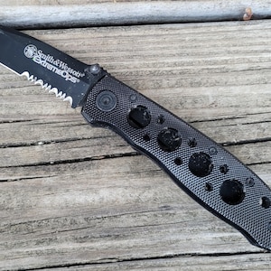 May include: Black Smith & Wesson Extreme Ops folding knife with a partially serrated blade. The handle features a textured grip with several circular cutouts. The knife is set against a weathered wooden surface.