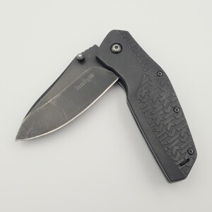 Op de afbeelding: Een zwart vouwmes met een gedeeltelijk open lemmet. Het lemmet heeft een donkere afwerking en de handgreep heeft een gestructureerd patroon. Het mes is een praktisch hulpmiddel voor diverse taken.