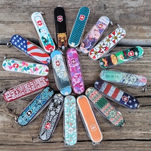 May include: A collection of Swiss Army knives with various designs, including the American flag, floral patterns, and a bear. The knives feature the Swiss cross logo and are arranged on a wooden surface. Each knife has a silver key ring.
