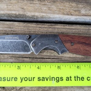 May include: A folding knife with a stainless steel blade and a wooden handle. The blade is marked with the word "BROWNING". The knife is placed on a wooden surface next to a ruler.