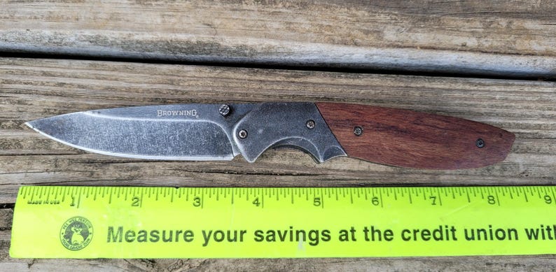 May include: A folding knife with a stainless steel blade and a wooden handle. The blade is marked with the word "BROWNING". The knife is placed on a wooden surface next to a ruler.