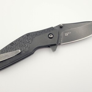 Op de afbeelding: Een zwart vouwmes met een getextureerde handgreep en een zilveren zakclip. Het lemmet is donkergrijs met de tekst "3850BLK CAT" erop gedrukt. Het mes is ontworpen voor dagelijks gebruik.