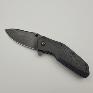 Op de afbeelding: Een zwart vouwmes met een getextureerde handgreep en een gedeeltelijk gekarteld lemmet. Het lemmet heeft de merknaam "Kershaw" gegraveerd. Het mes wordt getoond op een lichtgrijze achtergrond.