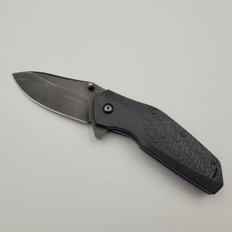 Op de afbeelding: Een zwart vouwmes met een getextureerde handgreep en een gedeeltelijk gekarteld lemmet. Het lemmet heeft de merknaam "Kershaw" gegraveerd. Het mes wordt getoond op een lichtgrijze achtergrond.