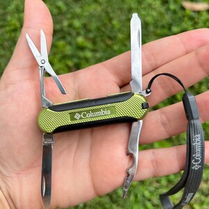 May include: A multi-tool with a lime green and black textured handle featuring the Columbia logo. The tool includes a small blade, scissors, a flathead screwdriver, and a bottle opener. A grey lanyard with the Columbia logo is attached to the side.