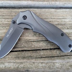 May include: A gray folding knife with the text "TAC-FORCE" on the blade. The knife is open, revealing a sharp blade. The handle has a curved design with black accents. The knife is lying on a wooden surface.