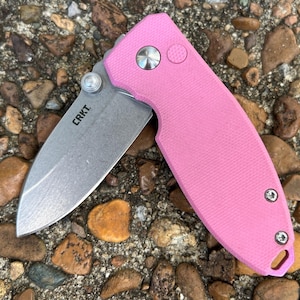 May include: A folding pocket knife with a bright pink textured handle and a silver-toned stainless steel blade. The blade features the CRKT logo printed in black near the pivot point. The handle includes a silver pivot screw and two small screws.