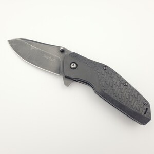 Op de afbeelding: Een zwart vouwmes met een getextureerde handgreep en een gedeeltelijk gekarteld lemmet. Het lemmet heeft een versleten uiterlijk. Het mes is gesloten en ligt op een wit oppervlak. De merknaam "Kershaw" is zichtbaar op het lemmet.