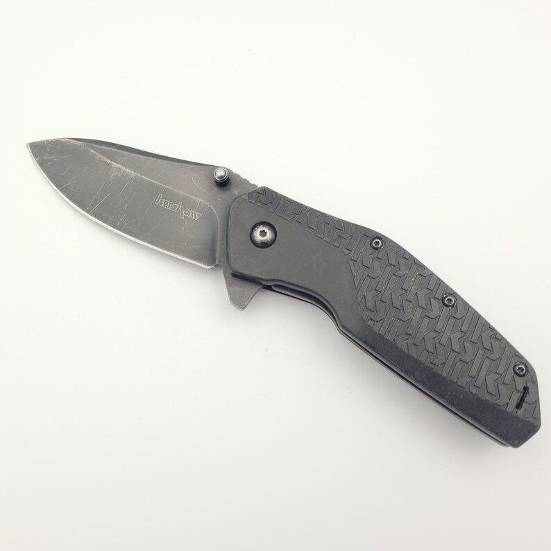 Op de afbeelding: Een zwart vouwmes met een getextureerde handgreep en een gedeeltelijk gekarteld lemmet. Het lemmet heeft een versleten uiterlijk. Het mes is gesloten en ligt op een wit oppervlak. De merknaam "Kershaw" is zichtbaar op het lemmet.