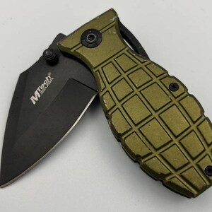 May include: A folding knife with a black blade and an olive green grenade-shaped handle. The blade is partially open, displaying the MTech USA logo. The handle features a textured, grid-like design.