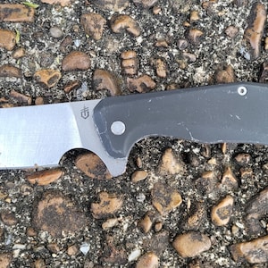 Könnte beinhalten: Ein silbernes und graues Klappmesser mit einer Klinge im Cleaver-Stil. Das Messer hat einen grauen Griff und liegt auf einem Bett aus kleinen Steinen. Die Klinge hat ein eingraviertes Logo.