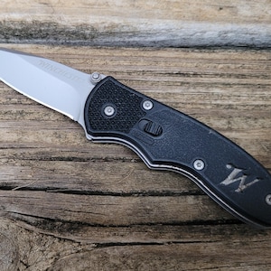 May include: A Winchester folding knife with a silver blade and a black handle. The blade is partially open, with the word "Winchester" visible. The handle has a textured grip and the letter "W".