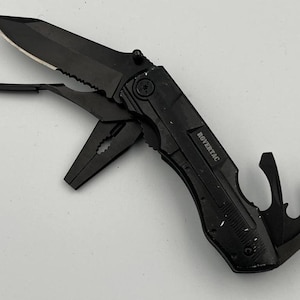 May include: A black multi-tool featuring a partially serrated folding blade, pliers, and various integrated implements. The handle is marked with the text ROVERTAC and displays a textured grip. The tool is constructed from dark metal components.