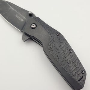 Op de afbeelding: Een zwart vouwmes met een gedeeltelijk gekarteld lemmet. Het mes heeft een getextureerde handgreep met een herhalend patroon en een zichtbaar draaipunt. Het lemmet is gemarkeerd met de merknaam "Kershaw".